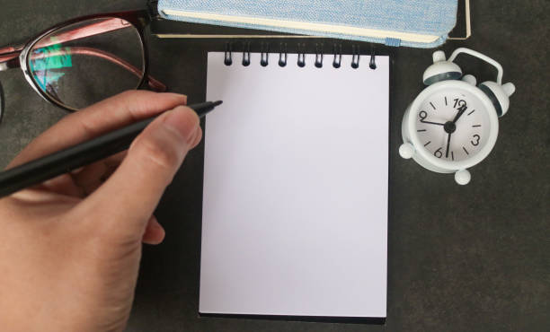 istock Hand writing in empty notebook for text on gray office desk table with books, pen, plant, eyeglass, clock and other. Blank notebook page for input the text in the middle. Top view, flat lay. 2191989001