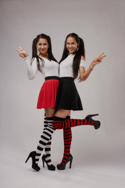 Two joyful young women wearing vibrant knee socks and stylish skirts are posing together, flashing peace signs and beaming smiles at the camera, radiating positivity and friendship