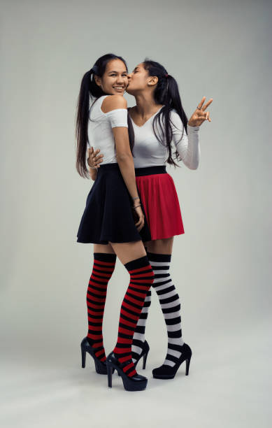 Two cheerful young women posing together in a studio, wearing colorful knee-high socks and high heels, sharing a kiss on the cheek while one shows a peace sign