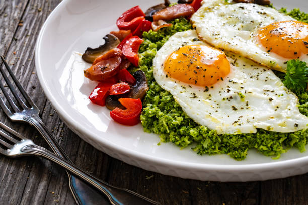 Breakfast - sunny side up eggs, grated broccoli, stir fried vegetables and sliced sausages served on wooden table