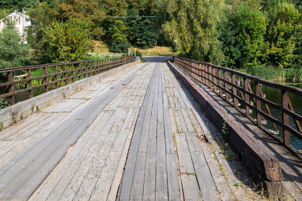 istock Old wooden bridge with railings over a river surrounded by lush green trees. 2192039980