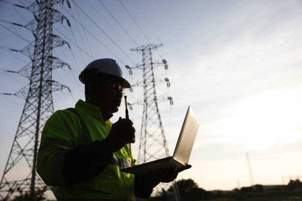 istock Senior high voltage engineer with green safety jacket and hardhat working on laptop and walkie talkie at the power plant under the electrical pole 2192057986