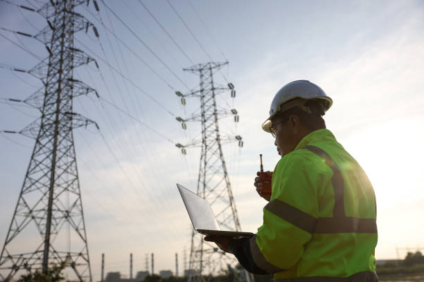 istock Senior high voltage engineer with green safety jacket and hardhat working on laptop and walkie talkie at the power plant under the electrical pole 2192058003