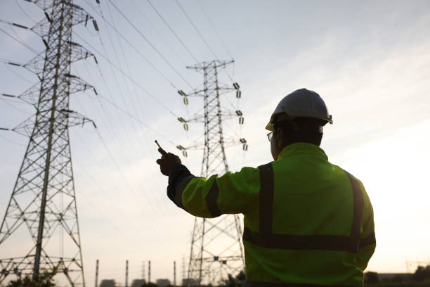 istock Senior high voltage engineer with green safety jacket and hardhat working on laptop and walkie talkie at the power plant under the electrical pole 2192058005