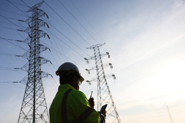 istock Senior high voltage engineer with green safety jacket and hardhat working on laptop and walkie talkie at the power plant under the electrical pole 2192058027