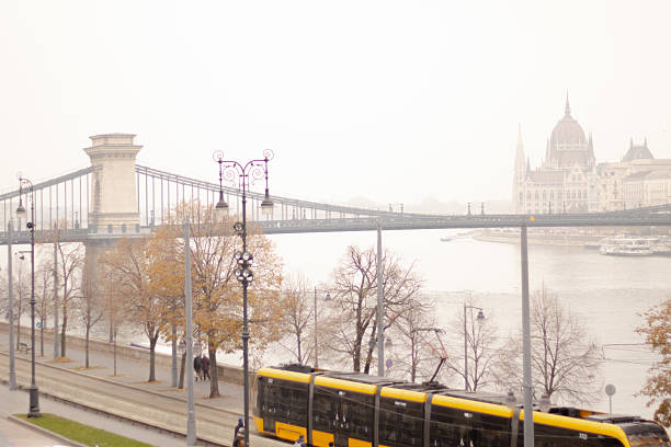 A yellow tram crosses while bridge enveloped in fog, with a faint view of a historic city in the background. The soft light creates a serene atmosphere.