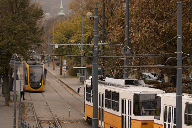 A tram moves along a tree-lined street in Budapest, surrounded by autumn leaves and urban scenery. The image captures daily life in the city during the fall season.