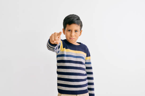 Boy wearing a striped sweater in blue, yellow, and gray, pointing directly at the camera with a serious expression and a determined gesture. White background emphasizes his action