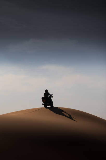 A solitary motorcyclist rides atop a desert dune, silhouetted against a vast, moody sky