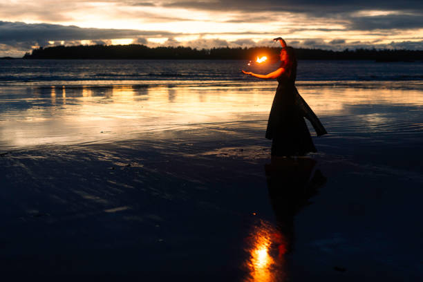 A colorfully dressed fire performer with rainbow mohawk punk hair, angel wings, and tribal body paint breathes fire from a torch during an outdoor festival at dusk, expressing bold creativity and energy.