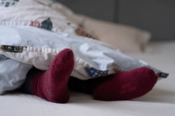 istock Close up of cozy maroon socks peeking out from under a soft, patterned duvet featuring a whimsical house print. The image conveys warmth and comfort, perfect for winter themes. 2192102525