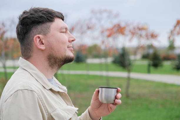 young man wearing yellow scarf enjoying autumn days breathing fresh air, enjoying walking in the park, drinking from thermos