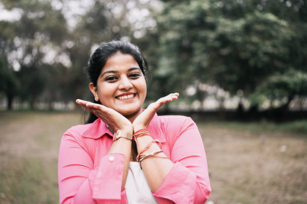 A vibrant young woman dressed in a bright pink shirt radiates happiness as she poses playfully with her hands framing her face. Set against the backdrop of a serene park, her wide smile and joyful demeanor embody positivity and carefree charm. Perfect for themes of self-confidence, outdoor lifestyle, and personal happiness