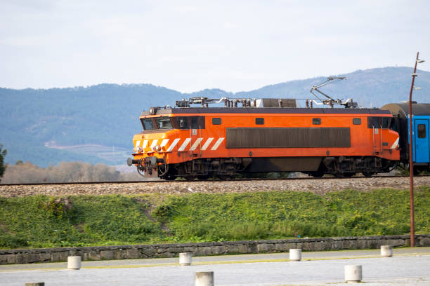 An electric train in vibrant orange color speeds through a picturesque landscape, showcasing modern transportation against a backdrop of rolling hills and greenery.