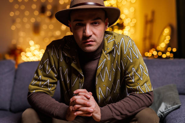A stylish man wearing a patterned jacket and hat sits pensively indoors, with warm yellow fairy lights in the background, creating a cozy and artistic atmosphere perfect for creative storytelling or editorial concepts.