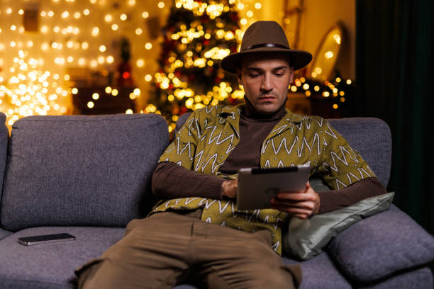 istock Man relaxing on couch using a tablet in a warmly lit festive room 2192125128