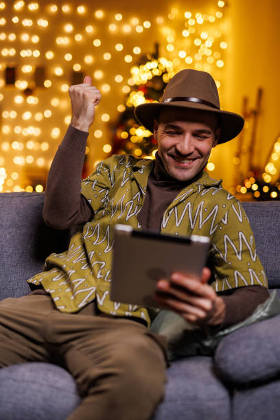 istock Man celebrating with a tablet while sitting on a festive holiday-decorated couch 2192125167