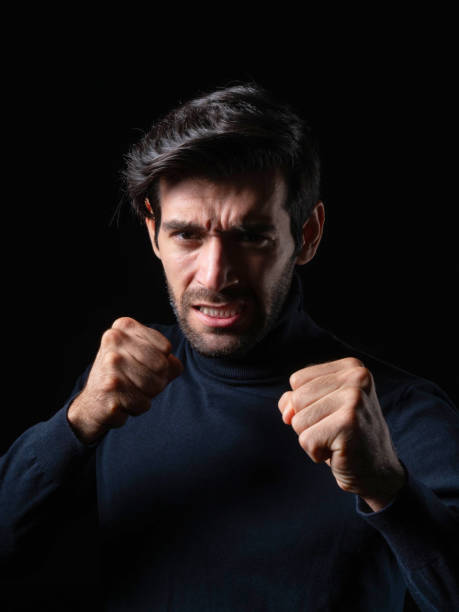istock Angry young man with an intense expression, clenching his fists in a defensive boxing pose, wearing a dark turtleneck sweater, isolated on a black background, symbolizing anger, determination 2192132210