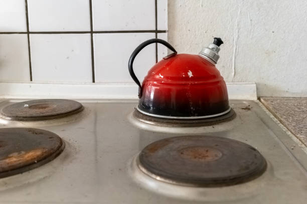 A bright red kettle sits on a stove, showing signs of use. The kitchen features a tiled wall and stainless steel burners, creating a warm atmosphere