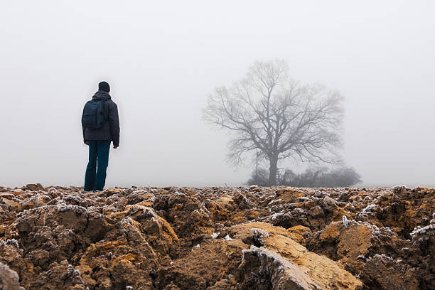 Man with backpack walking on frost field with leafless tree in fog