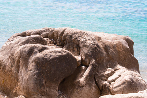 a large rock on the shore, next to clear blue water. The rock surface is uneven and has a variety of textures. The water looks clear and calm, shimmering in the light.