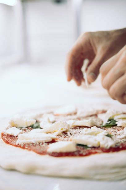 A close-up image of a freshly prepared Italian pizza being handcrafted by a chef. The dough is topped with a vibrant layer of tomato sauce, generous pieces of fresh mozzarella, and flavorful toppings like sausage crumbles. Captured in action, the hands of the chef delicately place the ingredients, emphasizing the artisanal process of pizza-making. The scene reflects the authenticity and care of traditional Italian cuisine, set against the clean and minimal backdrop of a marble work surface in a professional Italian restaurant.