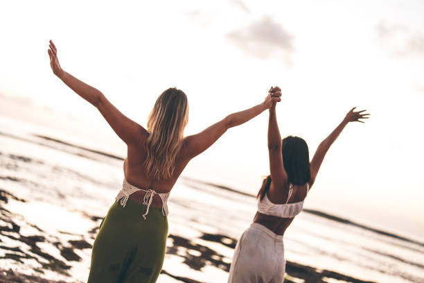 Two women with arms raised, standing on the beach during a serene sunset, symbolizing friendship, freedom, and joy in a natural and relaxing coastal environment.