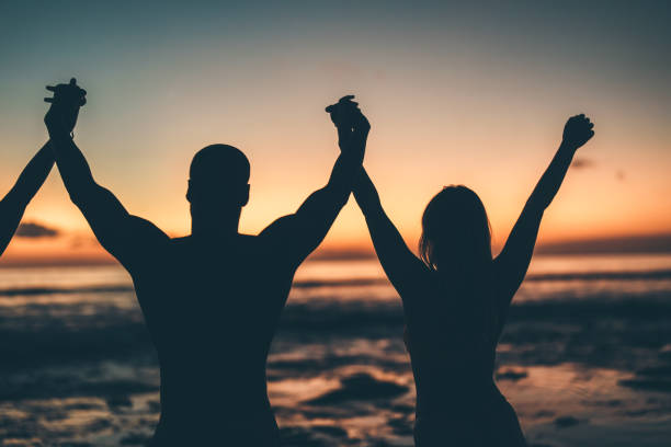 Silhouettes of individuals joyfully raising their hands against a vibrant sunset at the beach, evoking emotions of freedom, connection, and celebration amidst a serene and picturesque natural backdrop.