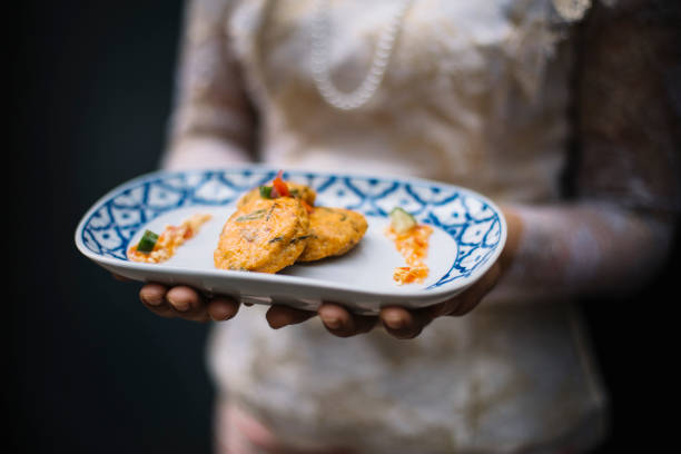 A beautifully plated serving of Tod Mun Pla, or Thai fish cakes, featuring golden-brown, deep-fried fish patties with aromatic herbs and spices. The dish is elegantly arranged on a traditional Thai blue and white ceramic plate, garnished with fresh vegetables and accompanied by a drizzle of sweet chili sauce. A server dressed in traditional Thai attire presents the dish, enhancing the cultural and authentic dining experience. The warm tones and soft lighting highlight the crispy texture and rich flavors of this classic Thai appetizer, making it a perfect representation of traditional Thai street food served in a fine dining setting.
