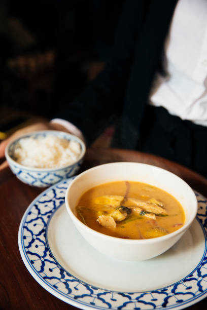 A beautifully presented serving of Thai Massaman curry, featuring a rich, coconut milk-based broth with tender meat, potatoes, and aromatic spices. The dish is elegantly served in a white ceramic bowl, placed on a traditional Thai blue and white patterned plate, alongside a separate bowl of fluffy jasmine rice. The warm tones of the curry contrast beautifully with the refined table setting, enhancing the luxurious and cultural dining experience. Massaman curry is a perfect combination of sweet, savory, and mildly spicy flavors, representing the depth and complexity of Thai cuisine.