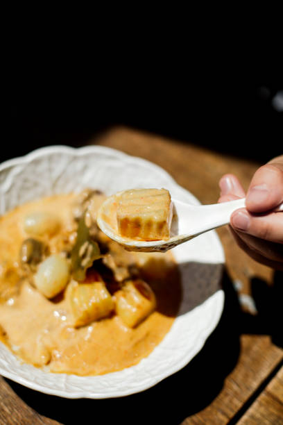 A beautifully presented bowl of Thai Massaman curry, a slow-cooked, coconut-based dish featuring tender chunks of beef, soft potatoes, pearl onions, and roasted peanuts in a thick, fragrant sauce. The dish is served in an elegant white ceramic bowl, highlighting its rich and creamy texture. A spoon lifts a piece of tender potato, capturing the velvety consistency of the sauce. The warm, golden hues of the curry contrast beautifully with the rustic wooden table, enhancing the depth of its flavors. This dish is a perfect example of Thai cuisine’s unique blend of aromatic spices and smooth coconut richness, offering a mild yet deeply flavorful experience.