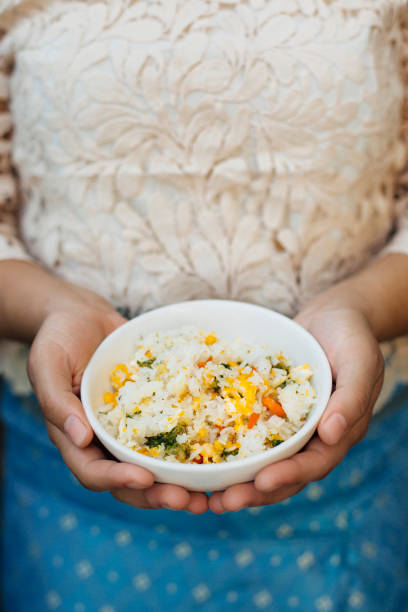 A bowl of freshly made Thai-style fried rice, featuring soft jasmine rice stir-fried with vibrant vegetables, scrambled eggs, and aromatic seasonings. The dish is held in the hands of a person dressed in elegant traditional Thai attire, adding an element of cultural authenticity. The close-up shot highlights the fluffy texture of the rice, the rich golden color of the eggs, and the bright hues of the vegetables, creating an inviting and appetizing presentation. This popular dish is a staple in Thai cuisine, known for its balance of flavors and comforting taste, commonly enjoyed in homes and street food markets alike.