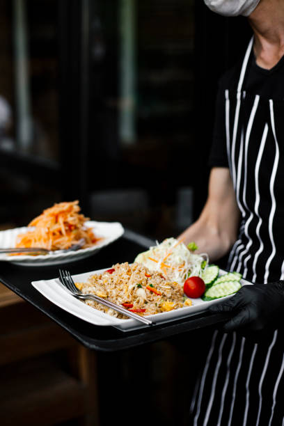 A restaurant server in a black and white striped apron carries a tray with a freshly prepared Thai meal, featuring fried rice with vegetables and a side of papaya salad. The dish is garnished with fresh cucumber slices, shredded cabbage, and cherry tomatoes, highlighting the vibrant colors of traditional Thai cuisine. The server wears black gloves, ensuring hygiene while serving customers. The background shows a casual dining setting, emphasizing the authenticity of the Thai culinary experience.