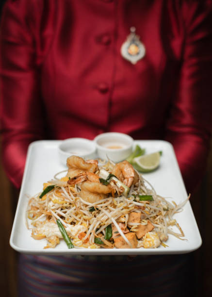 A beautifully presented plate of Pad Thai, Thailand's iconic stir-fried noodle dish, featuring rice noodles tossed with shrimp, chicken, egg, and fresh bean sprouts. The dish is garnished with chopped green onions and accompanied by a wedge of lime and a side of dipping sauce for extra flavor. Served on a clean white square plate, the meal is elegantly held by a server dressed in traditional Thai attire, adding cultural authenticity to the dining experience. The vibrant colors and rich textures of this dish highlight the balance of sweet, sour, salty, and umami flavors that define Thai cuisine.