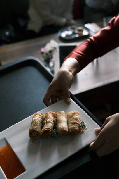 A close-up image capturing a hand serving a plate of crispy Thai spring rolls accompanied by a side of vibrant dipping sauce. The spring rolls are arranged neatly on a sleek white plate, garnished with fresh herbs, highlighting their golden texture. The background showcases a warm and inviting restaurant atmosphere, adding a sense of authenticity and cultural richness to this delightful appetizer. Perfectly encapsulates the essence of Thai dining and culinary presentation.