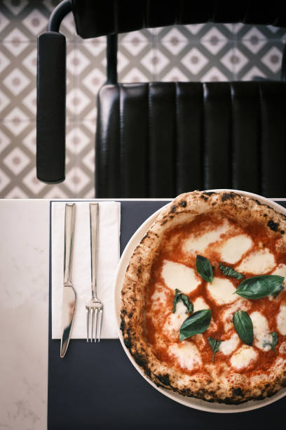 This image features a traditional Neapolitan Margherita pizza, placed on a modern dining setup with a black chair and geometric-patterned tile background. The pizza showcases its perfectly charred wood-fired crust, vibrant tomato sauce, creamy mozzarella, and fresh basil leaves. The elegant table setting, complete with a fork and knife on a clean napkin, highlights the refined yet cozy ambiance of the Italian restaurant. This visually appealing scene blends classic Italian culinary artistry with a contemporary dining atmosphere.