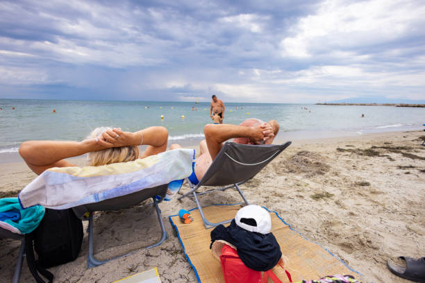 An elderly couple enjoys a serene day by the ocean, sunbathing on their beach chairs.\