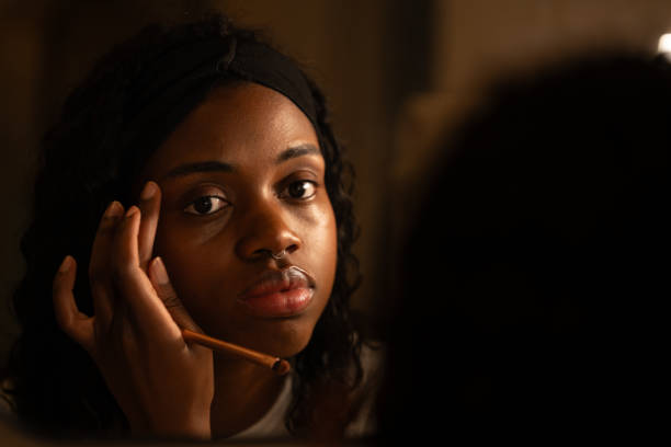 Woman applying makeup, gazing into mirror, soft lighting casting contemplative mood, highlighting personal beauty routine