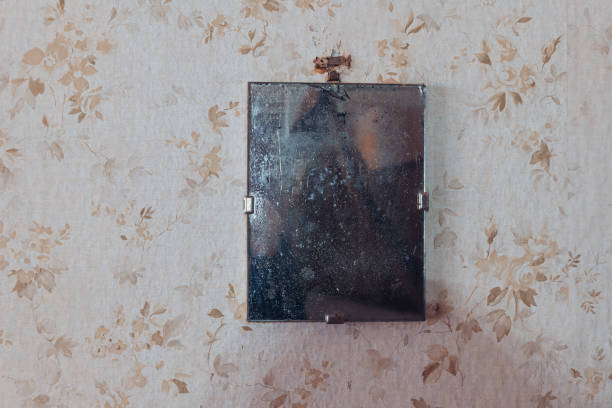 Reflective surface hangs on a floral patterned wall in a dimly lit room with dust accumulation stock photo