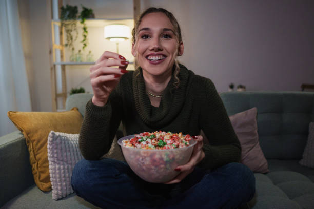 istock Young woman sitting on sofa eating popcorn and watching tv 2192191198