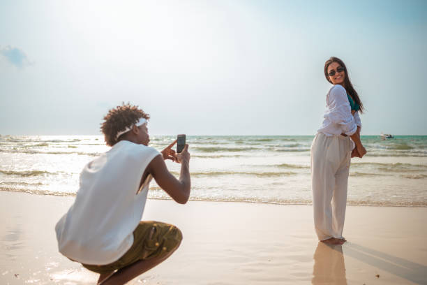 A content black male captures a joyful moment, photographing his radiant Hispanic girlfriend on a serene tropical beach, both dressed in casual summer attire.