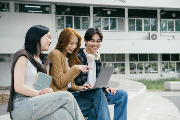 istock Three young adult students are sitting outside a university building, working together on a laptop and smiling, while one of them is pointing at the screen 2192204463