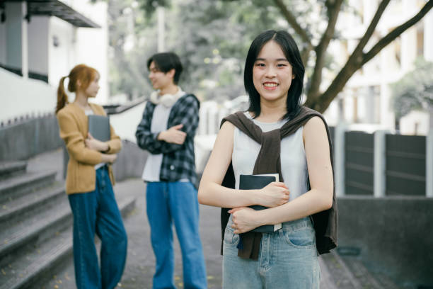 istock Smiling young female student holding books, posing confidently at university campus while friends chat and enjoy the vibrant atmosphere in the background 2192206288