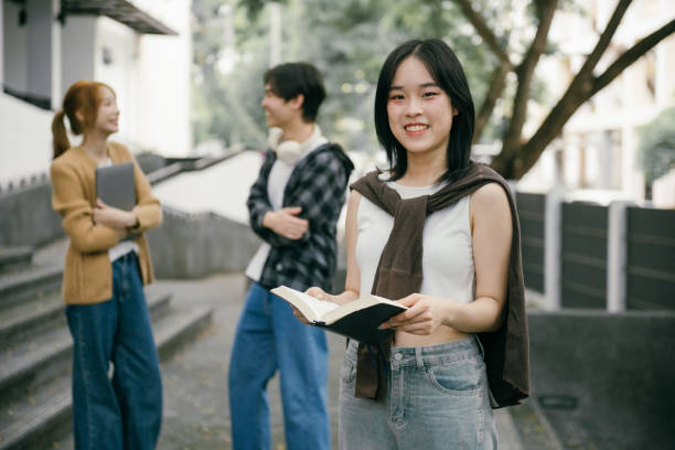 istock Young woman smiling at the camera while holding an open book, surrounded by two fellow students chatting in the background, enjoying a vibrant campus atmosphere 2192206395