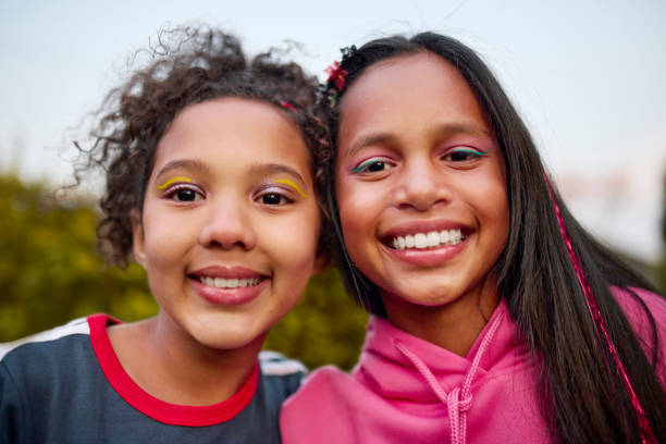 Two young girls with vibrant eye makeup smile brightly at the camera. One with curly hair, the other with straight hair and pink tinsel. A joyful, close-up portrait.