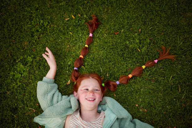 Overhead shot of a smiling red-haired girl with pigtail braids secured with colorful elastics, lying on green grass with one hand raised. Sunny outdoor scene.