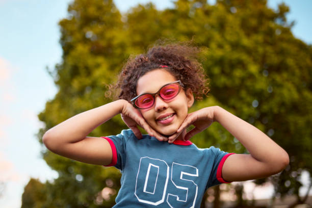 A happy young girl with curly hair and pink-tinted retro sunglasses smiles while playfully holding her face. She wears a blue '05' t-shirt against a blurred green and blue background.