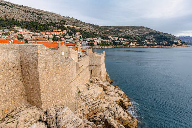 Dubrovnik, Croatia - January 13, 2019: Dubrovnik’s imposing stone walls hug the rocky cliffs along the Adriatic coast, showcasing the city’s strategic fortifications. The serene blue waters and surrounding landscape emphasize the harmonious balance between the city’s historic architecture and its natural coastal beauty.