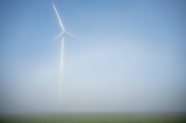 Wind turbines stand in the fog in a field with sunshine.