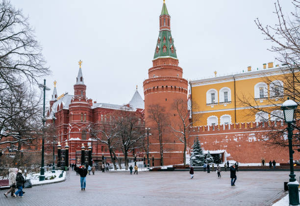 Kremlin tower rises above the red brick walls, framed by snowy trees and a clear winter sky. The open square features a few pedestrians walking, capturing the serene and historic winter scene.